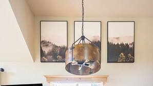 Et spiseområde med et træbord, fire stole, en hængende lampe, et fjernsyn og et vindue med udsigt til det fri.