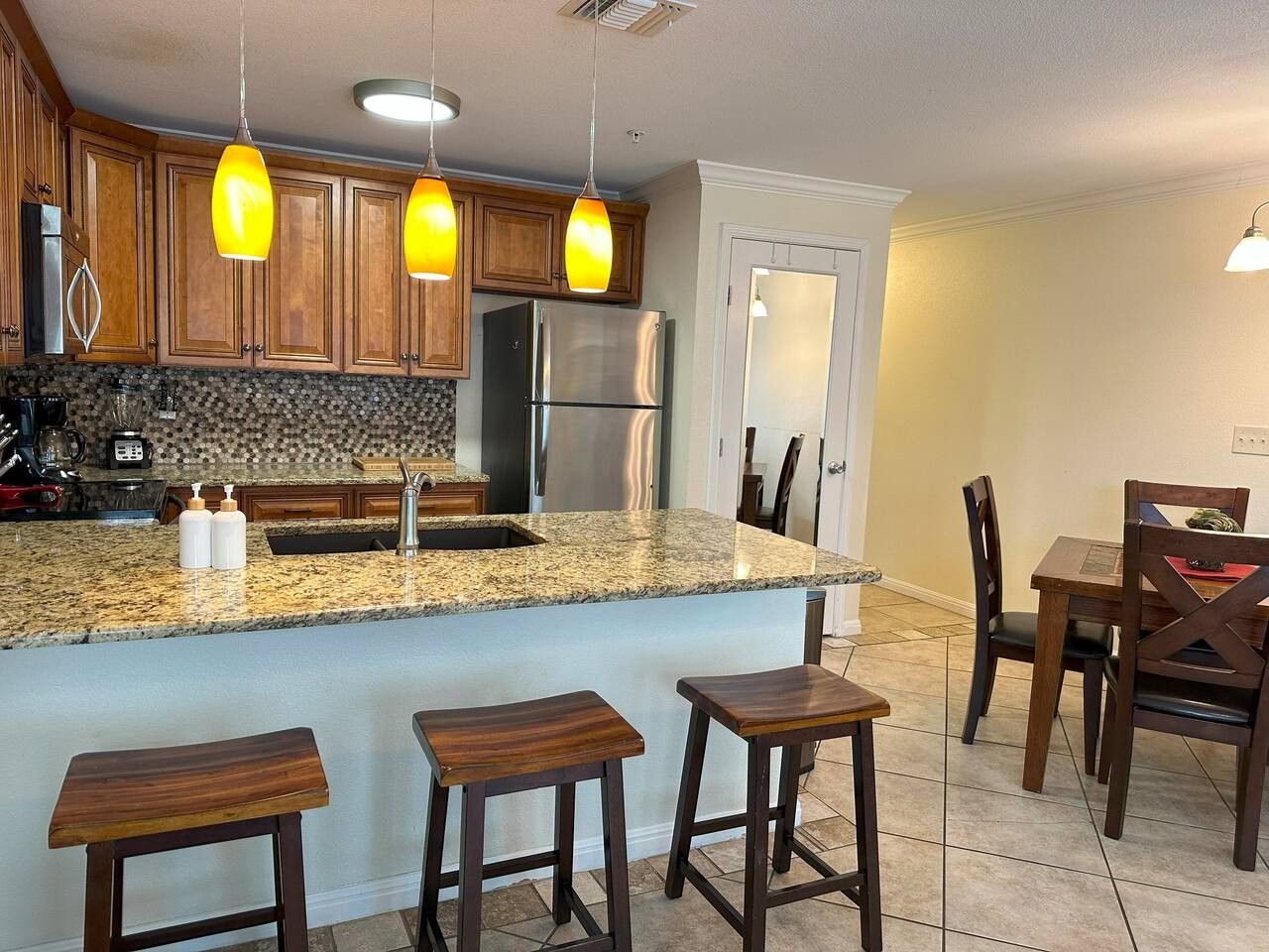 A kitchen with wooden cabinets, a granite countertop, a stainless steel refrigerator, and a bar area with stools.