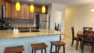 A kitchen with wooden cabinets, a granite countertop, a stainless steel refrigerator, and a bar area with stools.