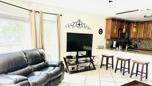 A living room with a leather sofa, a TV on a stand, a coffee table, and a kitchen area with wooden cabinets and a tiled backsplash. - The Blue Llama- on the Guadelupe river! 2 pools, 1 heated, 4 hot tubs (New Braunfels)