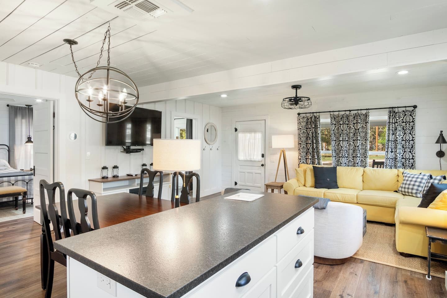 A modern kitchen with a central island, dining area with chairs, a sofa, and a TV mounted on the wall.