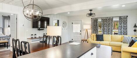 A modern kitchen with a central island, dining area with chairs, a sofa, and a TV mounted on the wall.