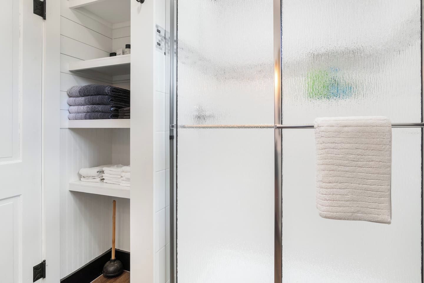 Une salle de bain avec une douche à parois de verre, une serviette blanche accrochée à la porte et des étagères avec des serviettes pliées.