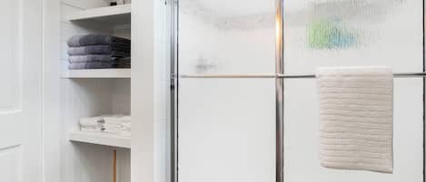 Une salle de bain avec une douche à parois de verre, une serviette blanche accrochée à la porte et des étagères avec des serviettes pliées.