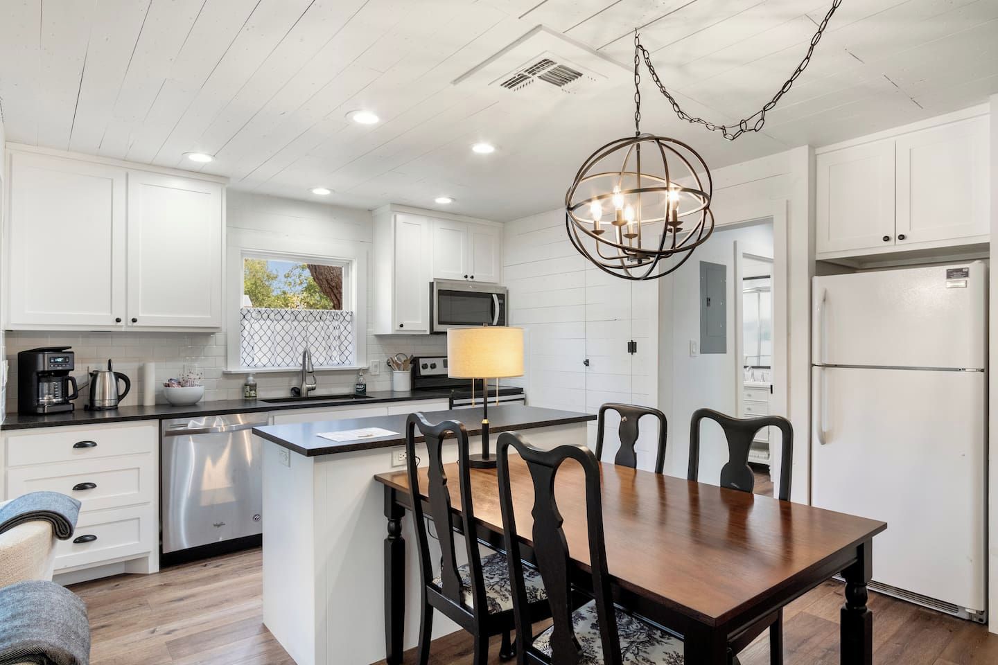 A modern kitchen with white cabinets, a stainless steel refrigerator, a dining table with chairs, and a pendant light.