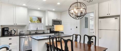 A modern kitchen with white cabinets, a stainless steel refrigerator, a dining table with chairs, and a pendant light.