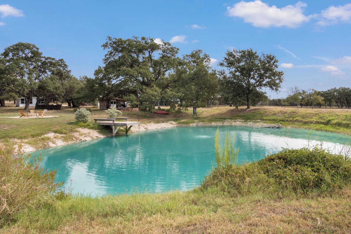 Una piscina exterior cristalina rodeada de césped y árboles, con una mesa de picnic y una pequeña construcción al fondo.