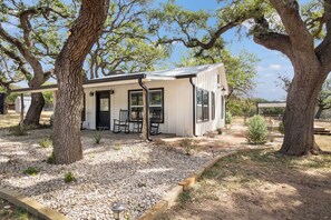 A modern house with a covered porch, surrounded by large trees and a gravel driveway. - Hill Country Mirage | 3BR Farmhouse w/ Pond & Bar (Bandera)