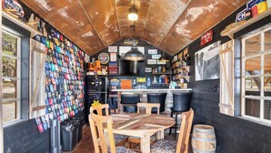 Um espaço de bar rústico com mesa e cadeiras de madeira, uma geladeira e uma parede decorada com diversos objetos de recordação.