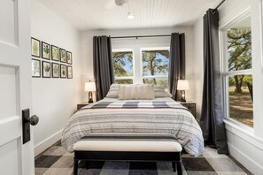 A bedroom with a bed, two bedside lamps, a bench, a window with curtains, and framed botanical prints on the wall.