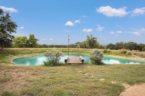 A tranquil outdoor swimming pool with a wooden deck and a flagpole, surrounded by grass and trees. - Hill Country Mirage | 3BR Farmhouse w/ Pond & Bar (Bandera)