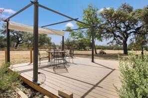 An outdoor patio with a table and chairs, surrounded by trees and a clear sky. - Hill Country Mirage | 3BR Farmhouse w/ Pond & Bar (Bandera)