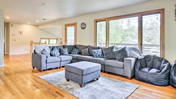 Amplia sala de estar con sofá modular, reposapiés y sillón de tina, ventanas grandes y piso de madera.