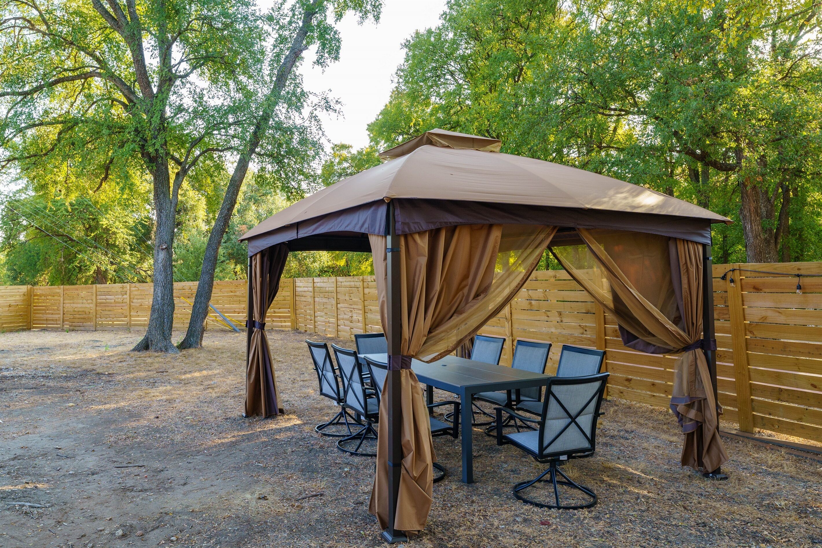 A covered outdoor seating area with a table and chairs, surrounded by trees and a wooden fence.
