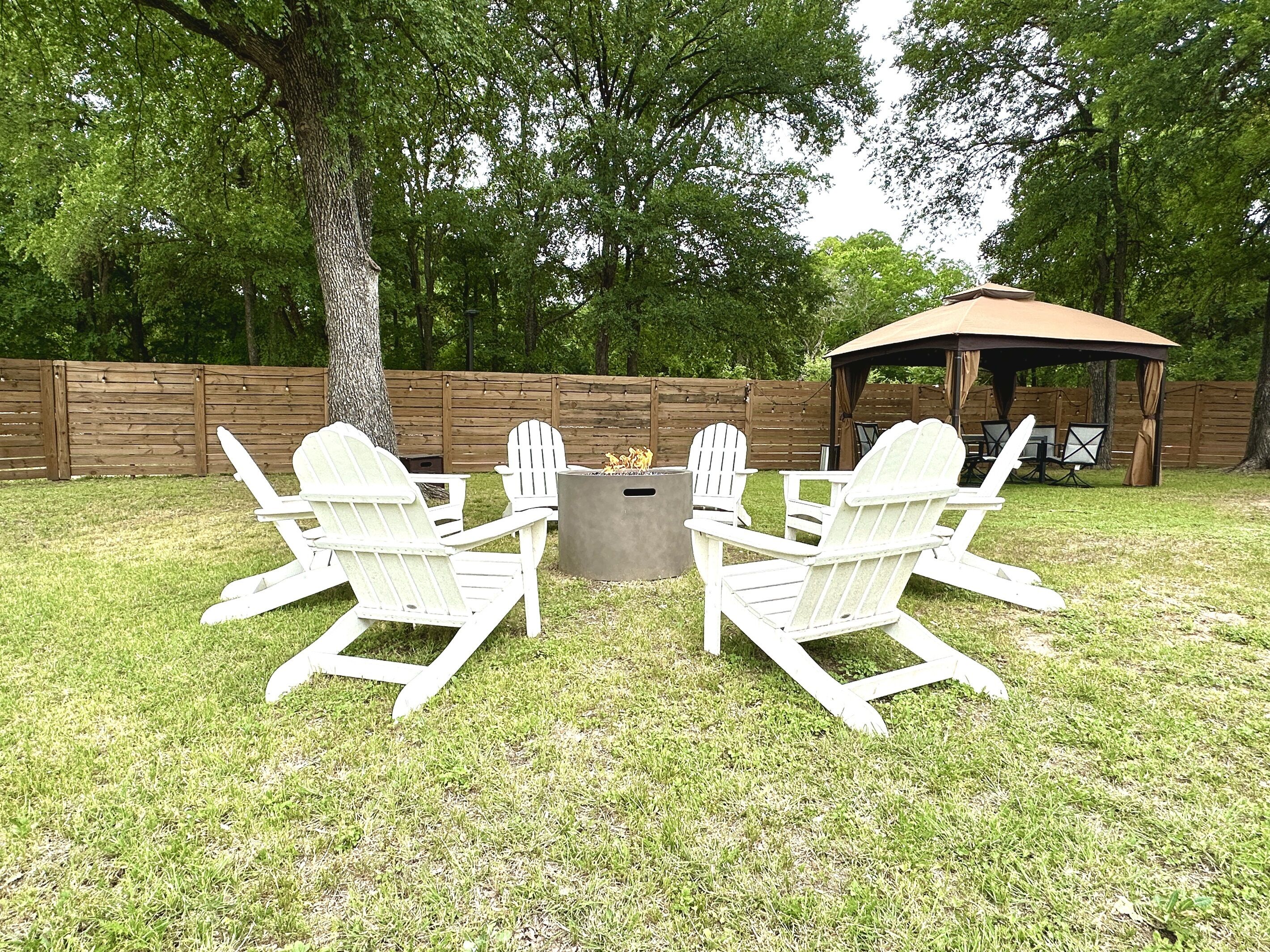 Un patio trasero con un brasero rodeado de sillas Adirondack, una valla de madera y un área de estar cubierta.