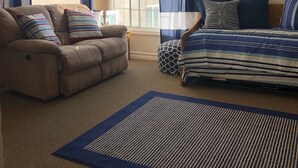 Una sala de estar con un sofá color beige, un sofá azul y blanco a rayas, y un reposapiés estampado. Se aprecia una ventana grande con cortinas azul y blanco, un cuadro con barcos de vela en la pared, y una alfombra azul y marrón.