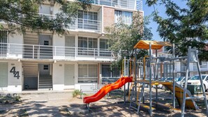 Une aire de jeux avec un toboggan rouge et des structures d’escalade, située devant un immeuble à plusieurs étages doté de balcons et d’un garage.