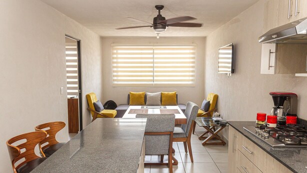 A modern kitchen with a dining area, featuring a granite countertop, wooden chairs, and a ceiling fan.