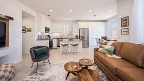 Una sala de estar moderna con un sofá marrón, una mesa de centro y un televisor. Se observa una cocina al fondo con armarios blancos y un refrigerador.