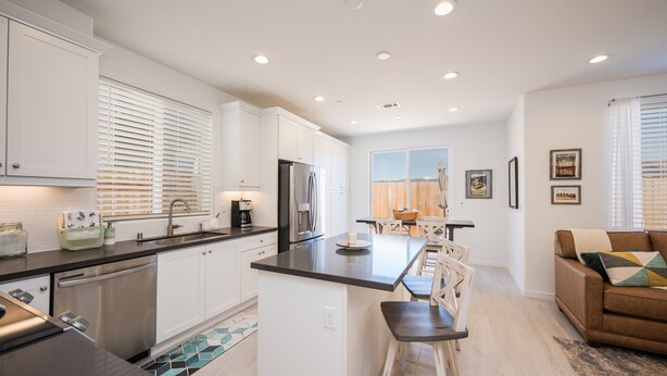 Una cocina moderna con armarios blancos, electrodomésticos de acero inoxidable y una isla central. Hay un comedor con mesa y sillas, y una sala de estar con sofá y una mesita de centro.