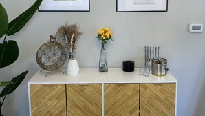 Ein modernes Wohnzimmer mit einem weißen Schrank, zwei gerahmten abstrakten Kunstwerken, einer Vase mit gelben Blumen und einem dekorativen Metallkorb.