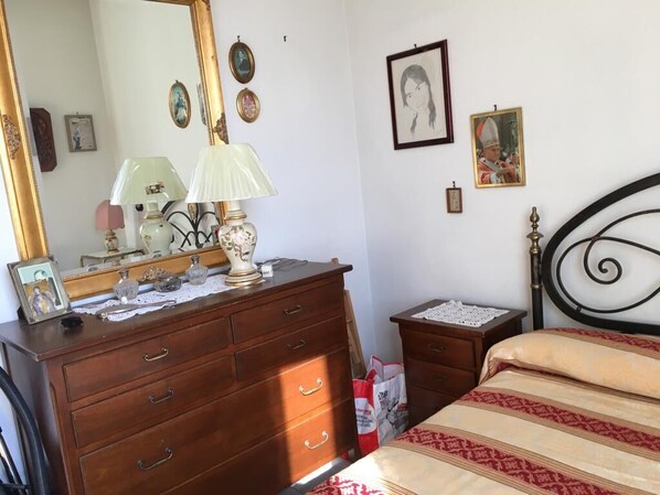 Une chambre à coucher avec une commode en bois, un lit recouvert d’une couvre-lit à motifs, une tête de lit en métal noir et une petite table de chevet avec une lampe.