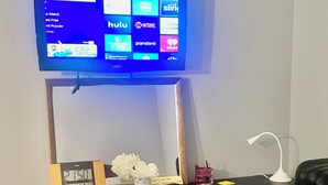 A television mounted on a wooden stand displaying a guest mode interface with various streaming services. Below the TV, there's a wooden dresser with drawers, a clock, and a small vase with flowers. A desk with a chair is positioned to the right.