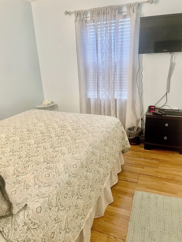 A bedroom with a bed, a nightstand, a dresser, a TV, and a window with curtains.