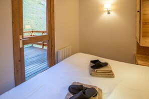 Quarto com porta de madeira que dá para uma varanda, cama com roupa de cama branca, um par de chinelos e luminária fixada na parede.