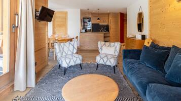 Un salon chaleureux avec une table basse ronde en bois, deux fauteuils à motifs, un canapé bleu foncé et, au fond, un coin cuisine.