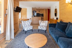 Uma sala de estar aconchegante com uma mesa de centro redonda de madeira, duas poltronas estampadas, um sofá azul-escuro e uma área de cozinha ao fundo.