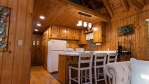 A wooden cabin interior with a kitchen area featuring a white refrigerator, a dining table with four chairs, and pendant lighting. - Hillside Haven - peaceful, private, pet friendly (Albany)