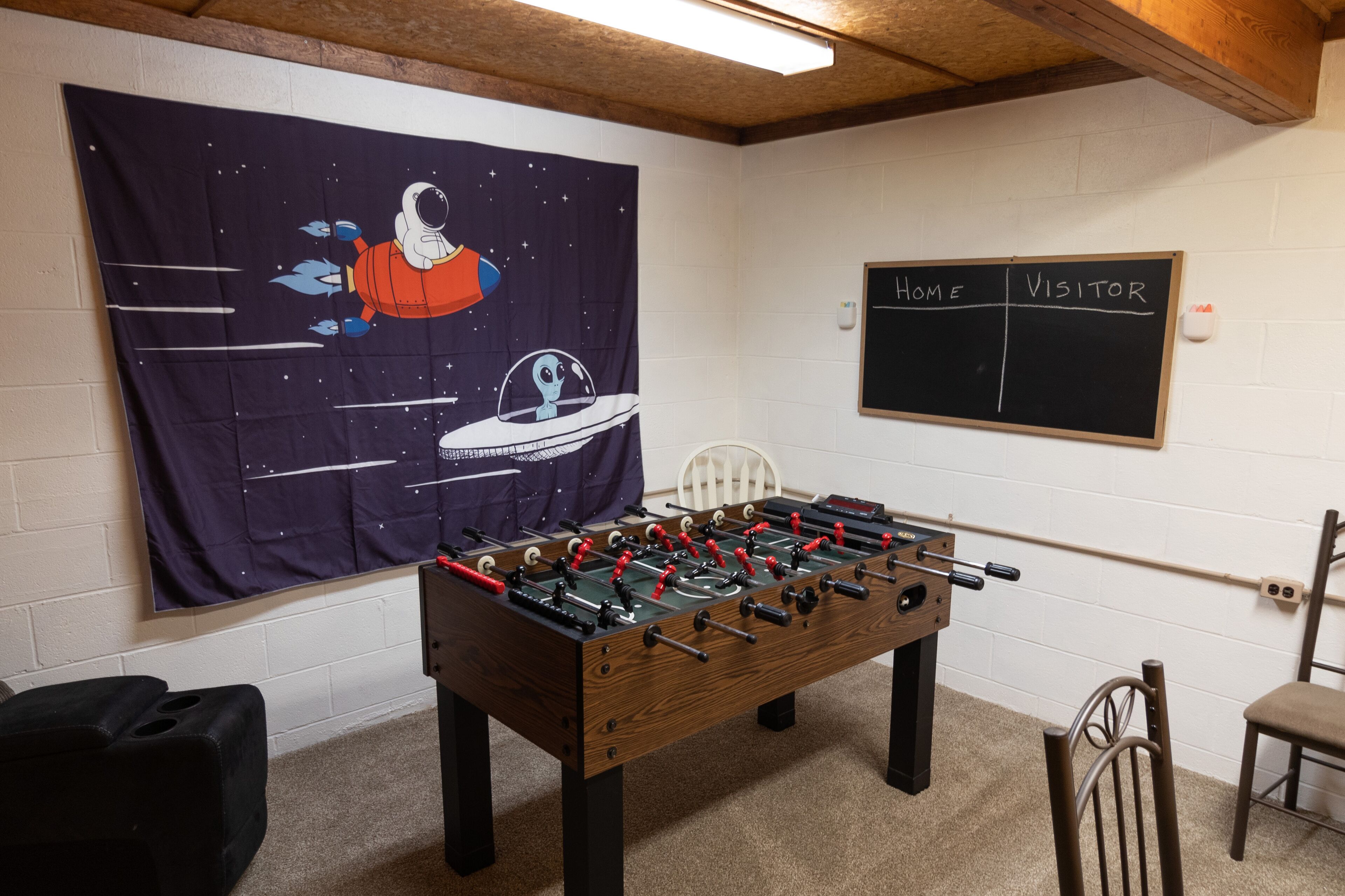 A room with a foosball table, a space-themed tapestry, and a chalkboard sign indicating 'HOME' and 'VISITOR' sides.