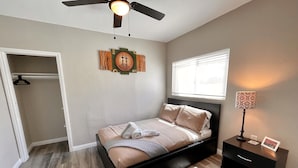 Ein Schlafzimmer mit Bett, Nachttisch, Deckenventilator, Fenster mit Jalousien und einem Wandbild.