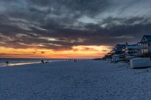 Une plage avec un coucher de soleil, un rivage sablonneux et des bâtiments situés en bord de mer.