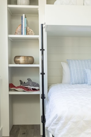Un lit superposé avec une tête de lit blanche, une étagère avec des livres et des objets décoratifs, et une table de chevet avec un coussin à rayures.