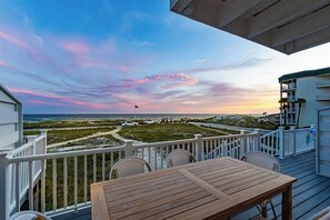 A wooden deck with a table and chairs, overlooking a beach with a clear sky and a flag.