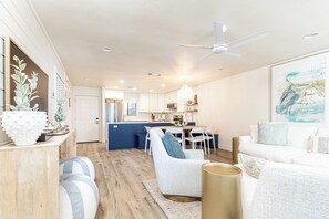 Un salon moderne avec un canapé, des fauteuils, une table basse et un coin repas, avec une cuisine en arrière-plan.