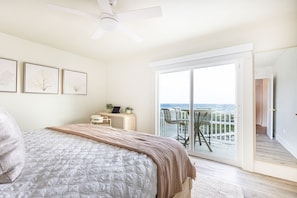 A bedroom with a bed, a desk, and a view of the ocean through a sliding glass door.