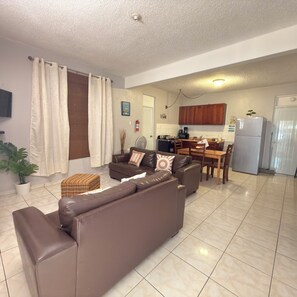 A living room with a brown sofa, a dining table, and a kitchen area with cabinets and appliances.