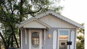 Une petite maison de plain-pied avec une porte blanche et des volets aux fenêtres, un chemin en béton menant à l’entrée et une boîte aux lettres sur le côté gauche.