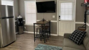 Un espace de vie compact comprenant un coin cuisine, un espace repas avec des tabourets, un téléviseur à écran plat fixé au mur et un canapé agrémenté d’un coussin décoratif.