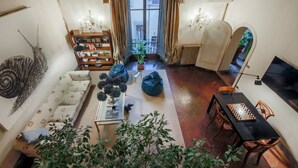 Uma sala de estar aconchegante com um sofá, uma mesa de centro com objetos decorativos, uma estante de livros, uma mesa de jantar com cadeiras e uma grande janela com cortinas.