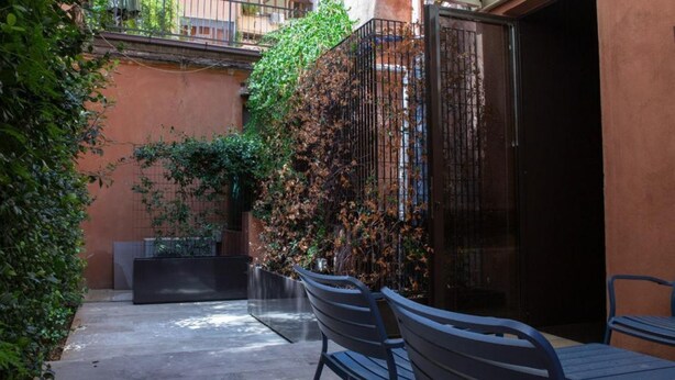 An outdoor patio with a concrete floor, metal chairs, a black planter, and a wall covered with climbing plants. - Luxury Suite Fori Imperiali (Roma)