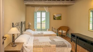 Un dormitorio con dos camas, una mesita de noche de madera con una lámpara, una silla de mimbre y un cuadro en la pared.