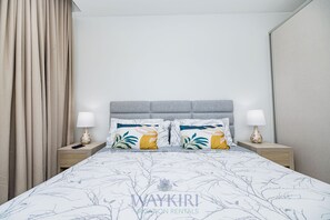 Una cama bien hecha con una colcha blanca y gris estampada, dos almohadas decorativas, un cabecero gris y dos mesitas de noche con lámparas.