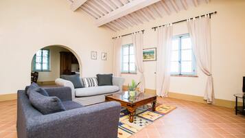 A living room with a sofa, armchairs, a coffee table, and a rug. There are two windows with curtains, a ceiling with wooden beams, and a door leading to another room.