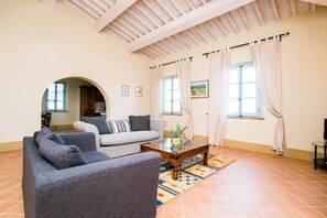 Un salon comprenant un canapé, des fauteuils, une table basse et un tapis. Il y a deux fenêtres avec des rideaux, un plafond avec des poutres en bois et une porte qui donne sur une autre pièce.