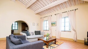 A living room with a sofa, armchairs, a coffee table, and a rug. There are two windows with curtains, a ceiling with wooden beams, and a door leading to another room.