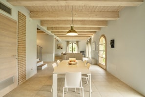 Un'ampia sala da pranzo con un lungo tavolo e sedie bianche, travi in legno al soffitto e grandi finestre che lasciano entrare la luce naturale.